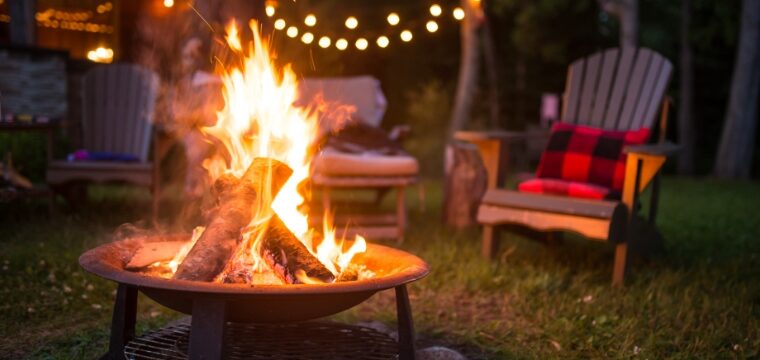 A campfire is burning in a backyard fire pit, with lawn chairs and lights in the background.