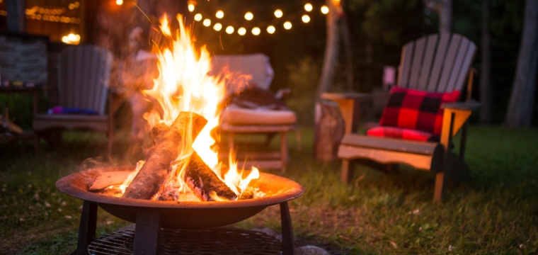 A campfire is burning in a backyard fire pit, with lawn chairs and lights in the background.