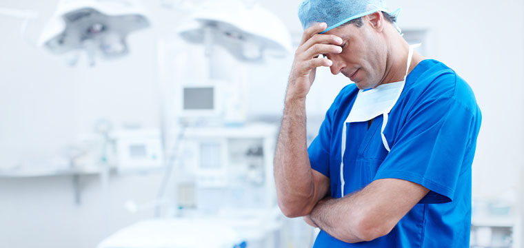 A male doctor in an operating room, holding his head due to negligence that led to medical malpractice.